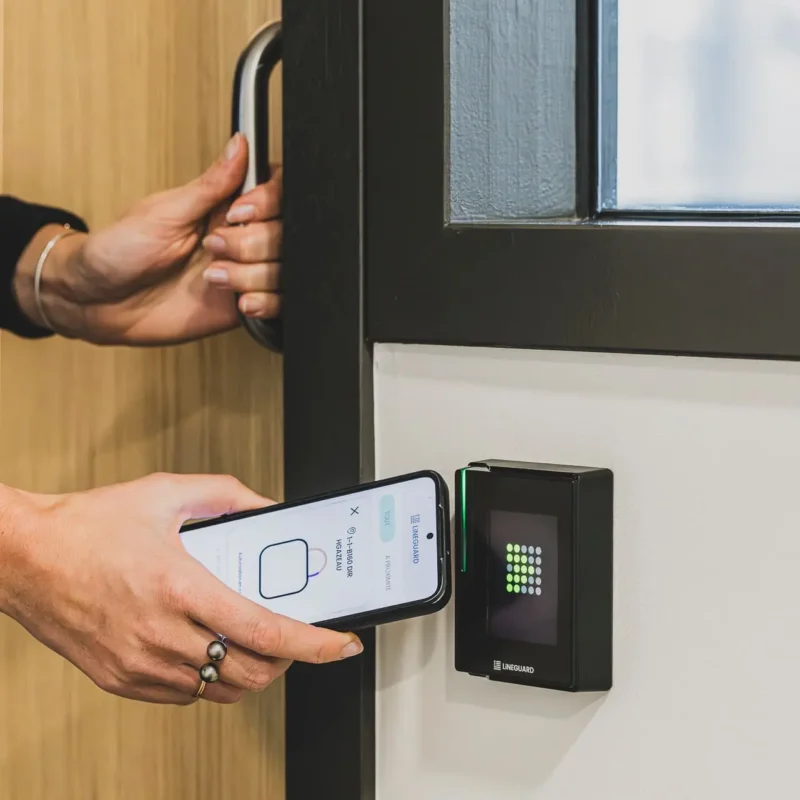 Une personne approche son smartphone de l'écran tactile d'un lecteur de badge mural noir marqué LiNEGUARD (modèle Line Ultra Touch).
