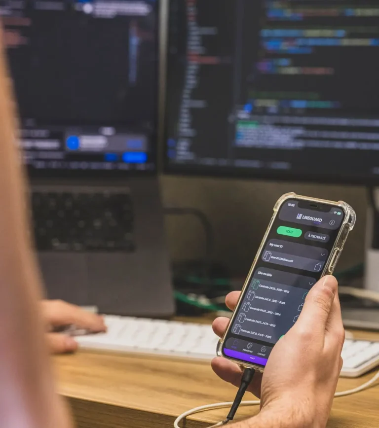 Zoom sur un smartphone apple affichant l'interface de l'application LiNEGUARD Access, tenu par un utilisateur devant deux écrans d'ordinateur affichant du code.