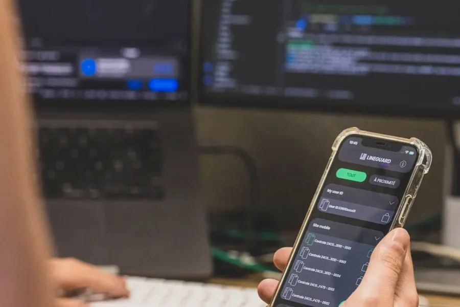 Close-up of an Apple smartphone displaying the LiNEGUARD Access app interface, held by a user in front of two computer screens showing code.