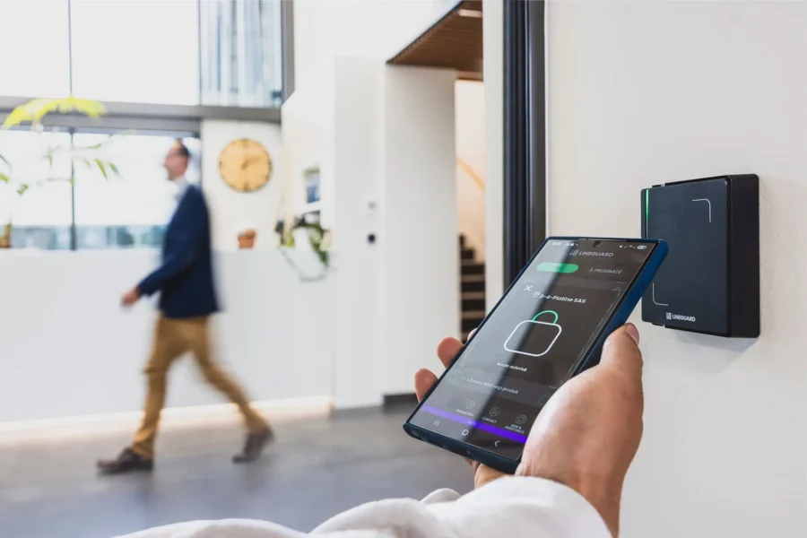 Vue en perspective dans le hall d'une entreprise où une main présente un smartphone devant un lecteur de badge mural noir (modèle Line Ultra Prox). Un employé est visible en arrière-plan flou.