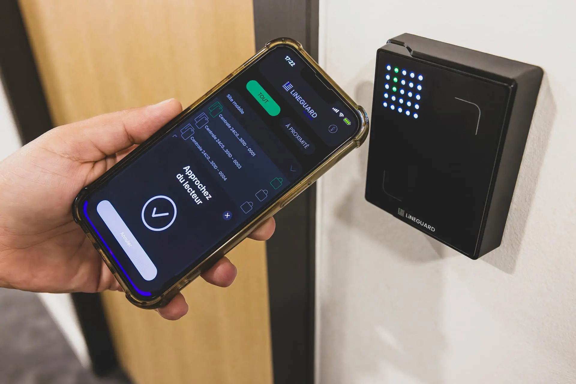 Un utilisateur présente son smartphone à l'application LiNEGUARD Access active devant un lecteur de badge mural noir rectangulaire avec des indicateurs LED colorés (modèle Matrix Ultra Prox).